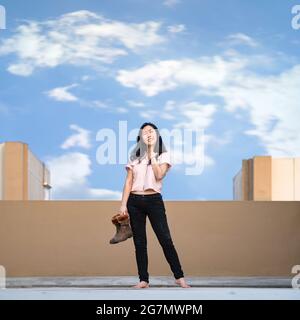 Asian woman stands and smiles alone with holds boots on rooftop in pastel blue - brown shade. Stock Photo