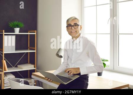 Happy senior businessman standing in office, holding laptop and looking at camera Stock Photo
