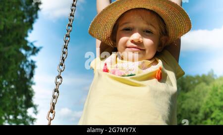 Portrait of a girl in a hat close-up on a background of blue sky. A beautiful girl in a yellow T-shirt is riding a swing. The concept of a happy caref Stock Photo