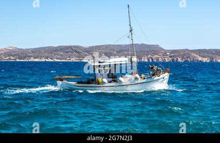 Cyclades, Greece. Fishing boat white traditional wooden vessel sailing in rough wavy sea. Rocky land, Greek island coast and clear blue sky background Stock Photo