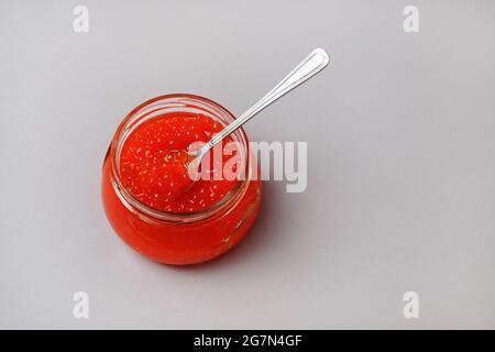 Red caviar in glass jar with spoon on gray background. Stock Photo