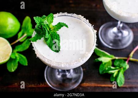 Coconut Mint Margaritas on a Dark Wood Background: Coconut and mint margaritas garnished with mint served in glasses rimmed with sugar and coconut Stock Photo