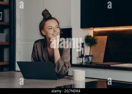 Young happy freelancer lady in cozy pajamas working from home on laptop Stock Photo