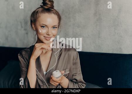 Attractive young female model in brown silk pajama sitting on bed with face cream Stock Photo