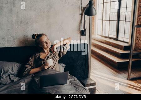 Happy relaxed positive young girl freelancer drinking coffee before to start working remotely at home Stock Photo
