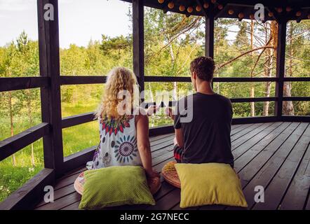 Young couple sit on pillows on wooden house patio, toasting celebrating holiday in wild nature in forest, glamping concept. Alone time together. Stock Photo