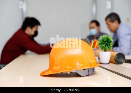 Close up image of a helmet on the desk with architects wearing protective masks working together in the backward, an architectural concept. Stock Photo
