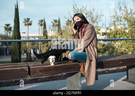 young woman with mask talking on the phone sitting on a bench. She wears black high boots and has a coffee cup leaning on the bench Stock Photo