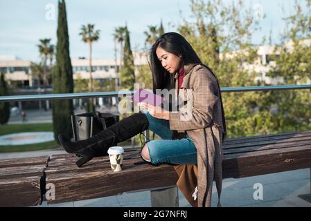young woman reading an e-book sitting on a bench while drinking some coffee Stock Photo