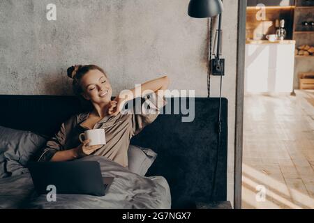 Happy beautiful female with closed eyes drinking coffee in bed before remote work at home Stock Photo