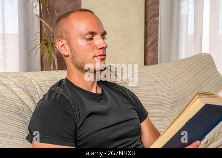 Young man reading a book in his living room sitting on the couch. Study concept. Stock Photo