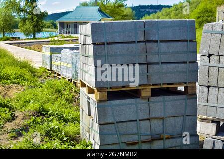 Pallets with gray paving slabs in park area on river bank. Plastic packaging protects the tile from atmospheric effects. Preparation for installation. Stock Photo