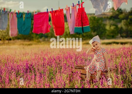 Little sweet baby sitting on wooden box in lavender field and posing for camera. Colorful clothes hanging on rope in field Stock Photo