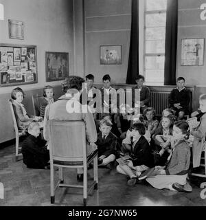 1950s, historical,  school, in the corner of a classroom, a female teacher sitting on a chair, reading a story to a group of young school school children, most of whom are sitting on the wooden parquet floor, England, UK. Stock Photo