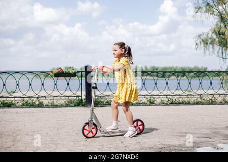 a happy, smiling girl in a dress rides a scooter along the river embankment in the city, a girl learns to ride a scooter on a sunny summer day Stock Photo