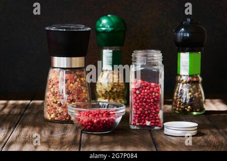Glass jar of pink pepper and various spices in glass mills on a dark wooden table close up Stock Photo
