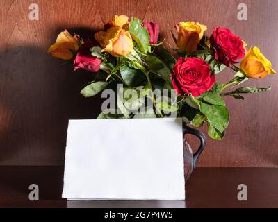 blank sheet of paper and red and yellow roses in ceramic jug on dark brown wooden background Stock Photo