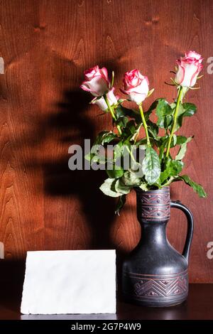 blank sheet of paper and pink roses in ceramic jug on dark brown wooden background Stock Photo