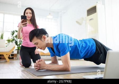 Smiling young woman watching  smartphone and man exercising and doing plank at home. Stock Photo