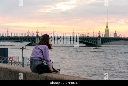 Young woman sitting on granite embankment, enjoying sunset view behind Troitskiy bridge on Neva river, during white night, St Petersburg, Russia Stock Photo
