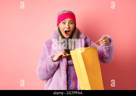 Shopping and fashion concept. Surprised asian grandmother open shop bag and looking amazed at camera, receive mother day gift, pink background Stock Photo