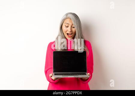 E-commerce concept. Amazed asian grandmother with grey hair, checking out promo online, showing laptop black screen, standing over white background Stock Photo