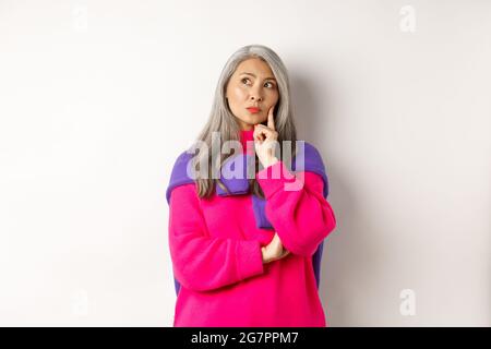 Thoughtful asian middle-aged mother looking upper left corner, thinking with serious face, standing over white background Stock Photo