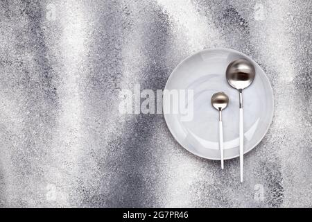 Empty clean ceramics plate and unused teaspoon with tablespoon. Crockery and cutlery on a gray grunge table, serving concept. Monochrome design menu. Stock Photo