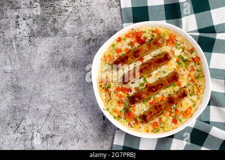 Oven baked rice with sausages and vegetables in a white baking dish on a dark grey background. Top view, flat lay Stock Photo