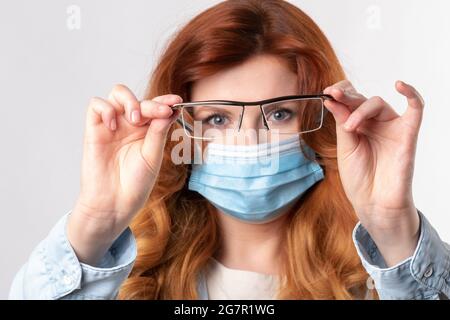 Attravtive red hair woman holds her glasses wearing antivirus protection mask for covid-19 epidemic virus Stock Photo