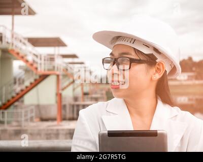 Female engineer in a white helmet talking radio communication (walkie talkie) and holding a digital tablet, stands in construction site of the hydroel Stock Photo