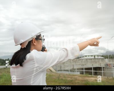 Female engineer in a white helmet using radio communication with construction worker at the dam construction site to generate electricity. Stock Photo