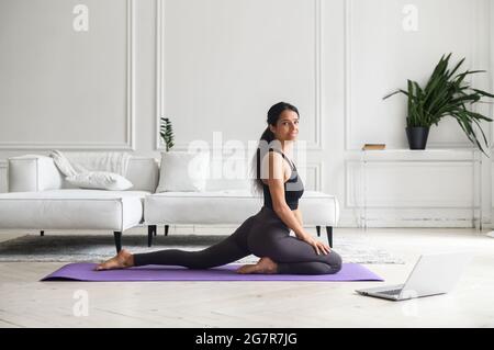 Young charming indian ethnicity female enjoys practicing yoga at home, doing asanas in front of the laptop. Mixed-race woman having personal fitness class online, look at screen and repeats exercises Stock Photo