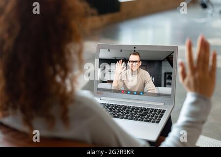 Young woman with curly hairstyle sitting at the laptop, chatting with male friend, colleague, talking about the news, work, new project, meeting online, making video call to man, waving Stock Photo