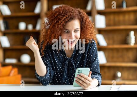 Surprised curly red-haired young woman using smartphone indoors, sitting at the desk in the office. Amazed happy lady reading message and rejoices, received good news, win online, raising fist up Stock Photo