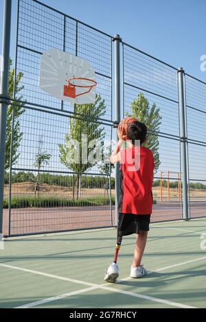 Back view of a basketball kid player with a leg prosthesis throwing the ball to the basket. Shooting basketball. Stock Photo