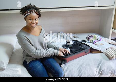 Portrait of a young black woman with afro hairstyle preparing clothes packing stuff in suitcase leaving for holidays trip in a room. Lifestyle concept Stock Photo