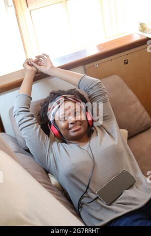 Portrait of a young black woman listening music with headphones indoor lying on a sofa.  Lifestyle concept. Stock Photo