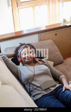 Portrait of a young black woman listening music with headphones indoor lying on a sofa.  Lifestyle concept. Stock Photo