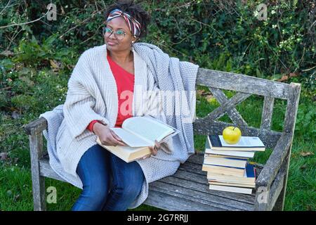 Portrait of a young black woman with afro hairstyle and glasses reading a book sitting on a garden old bench with a pile of books. Lifestyle concept. Stock Photo