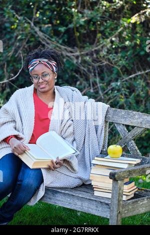 Portrait of a young black woman with afro hairstyle and glasses reading a book sitting on a garden old bench with a pile of books. Lifestyle concept. Stock Photo