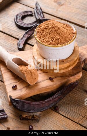 Dry carob pods and carob powder in a bowl over wooden background. Organic healthy ingredient for vegan vegetarian food and drinks, close up Stock Photo