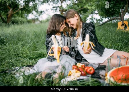 Two positive girls in stylish clothes sitting face to face on nature and holding banana in hands. Cute sisters having summer picnic on fresh air. Concept of family and happiness. Stock Photo