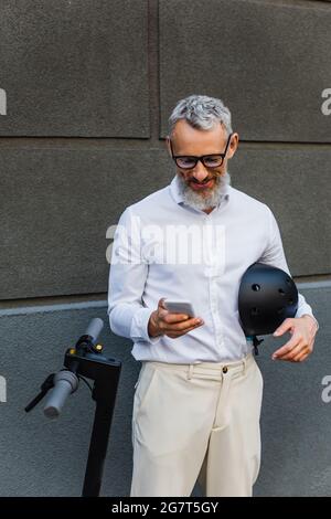 smiling and mature man in shirt holding helmet and using smartphone near electric scooter Stock Photo