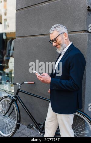 happy mature man in glasses using smartphone near bicycle on urban street Stock Photo