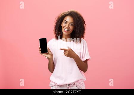 Suggest this model of phone. Happy charming african american curly-haired woman in stylish outfit holding smarpthone and pointing at device screen Stock Photo