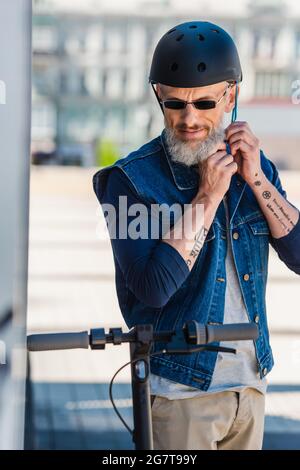 tattooed middle aged man in sunglasses wearing helmet near electric scooter Stock Photo