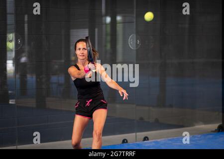 Woman playing padel in a blue grass padel court indoor - Young sporty woman padel player hitting ball with a racket Stock Photo