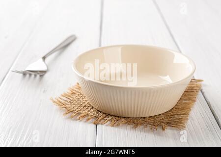 Empty beige bowl and metal fork on a white wooden table. Clean porcelain soup bowl on a table. Empty ceramic tableware ramekin for food design mockup. Stock Photo