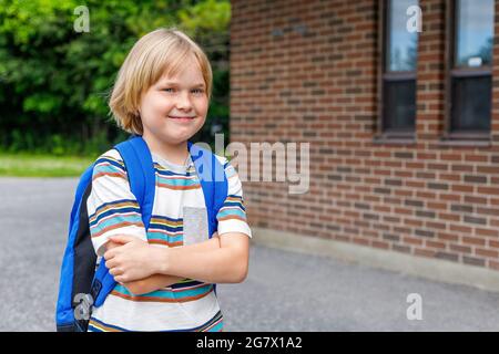 Beautiful happy child near school building at the schoolyard. Little blond student carrying blue backpack. Back to school concept. Stock Photo